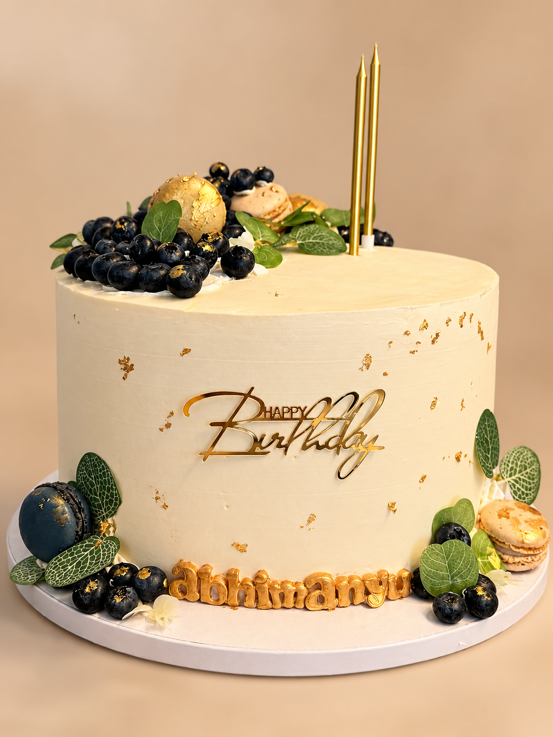 Tall ivory birthday cake with a gold Happy Birthday topper, gold-painted name, blueberries with gold leaf and macarons